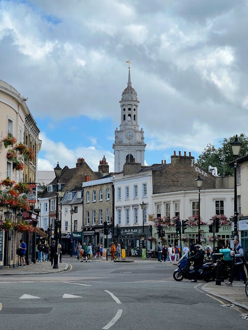 A lively street scene in Isleworth showing a variety of floral arrangements and hanging flower baskets decorating the shopfronts on the left side. In the foreground, a bouquet of fresh flowers with vi
