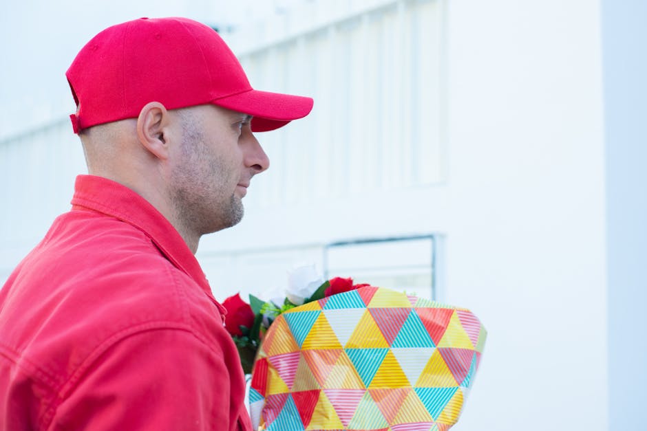 A delivery person wearing a red cap and red jacket holding a colorful floral bouquet wrapped in vibrant, geometric-patterned paper and tied with a red ribbon. The bouquet features a mix of fresh flowe