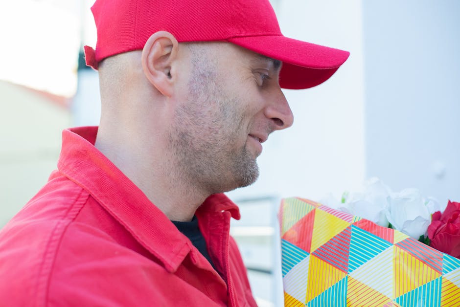 A man wearing a red cap and matching red jacket is holding a colorful bouquet of flowers wrapped in geometric patterned paper, with white and pink blooms visible. The flowers appear fresh with vibrant