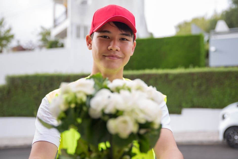 A delivery person wearing a red cap and yellow high-visibility vest is smiling and holding a bouquet of fresh white flowers, including roses and possibly peonies or ranunculus, wrapped in clear cellop