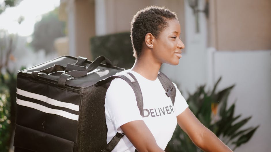 A professional female delivery courier with short curly hair, wearing a white T-shirt with the word 'DELIVER' printed on it, carrying a large black insulated insulated bag with white stripes on her ba