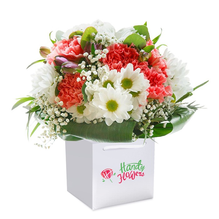 Resting gracefully in a crisp white square gift bag, this lush bouquet exudes timeless Islaeworth charm. Coral and peach-hued carnations, their frilled petals creating abundant, inviting texture, mingle beautifully with bright white daisies whose lime-green centers gleam with vibrancy. Sprigs of white baby's breath weave through, lending a sense of gentle movement and lightness, much like the blooms swaying near Isleworth Riverside Walk. Discreet buds tinged in lilac and rose rise to the left, hinting at new life and depth within the composition. Framing the entire arrangement is a generous band of deep green foliage, its broad, glossy leaves twisted and layered to give both structure and natural ease, mirroring the gardens of Syon Lane or Twickenham Road. The gift bag is adorned with a subtle floral emblem, its discrete elegance a nod to the high standards of local floristry. The bouquet's full, rounded form demonstrates expert technique, each flower carefully chosen and positioned for balance and harmony. Against a pure white background, the image glows with freshness and possibility. Such a bouquet is ideal for commemorating birthdays, anniversaries, or as an uplifting gesture for friends and loved ones throughout the Isleworth community, encapsulating the area's gentle vibrancy and spirit.
